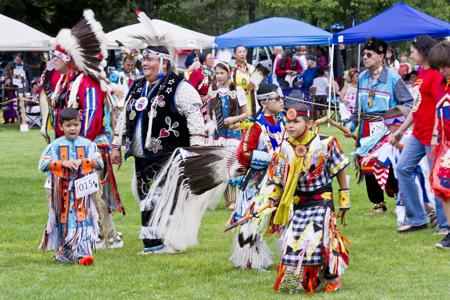 Image from a tribal pow wow with several adult and children dancing in native dance attire.