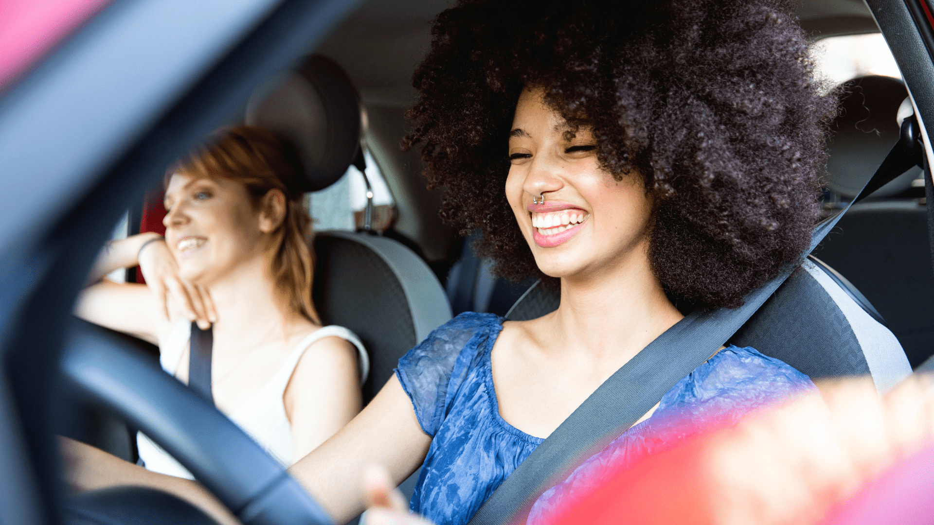 Photo of a young woman driving a car. She is smiling and wearing a seatbelt. The passenger is also a woman, she is smiling and wearing a seatbelt. 