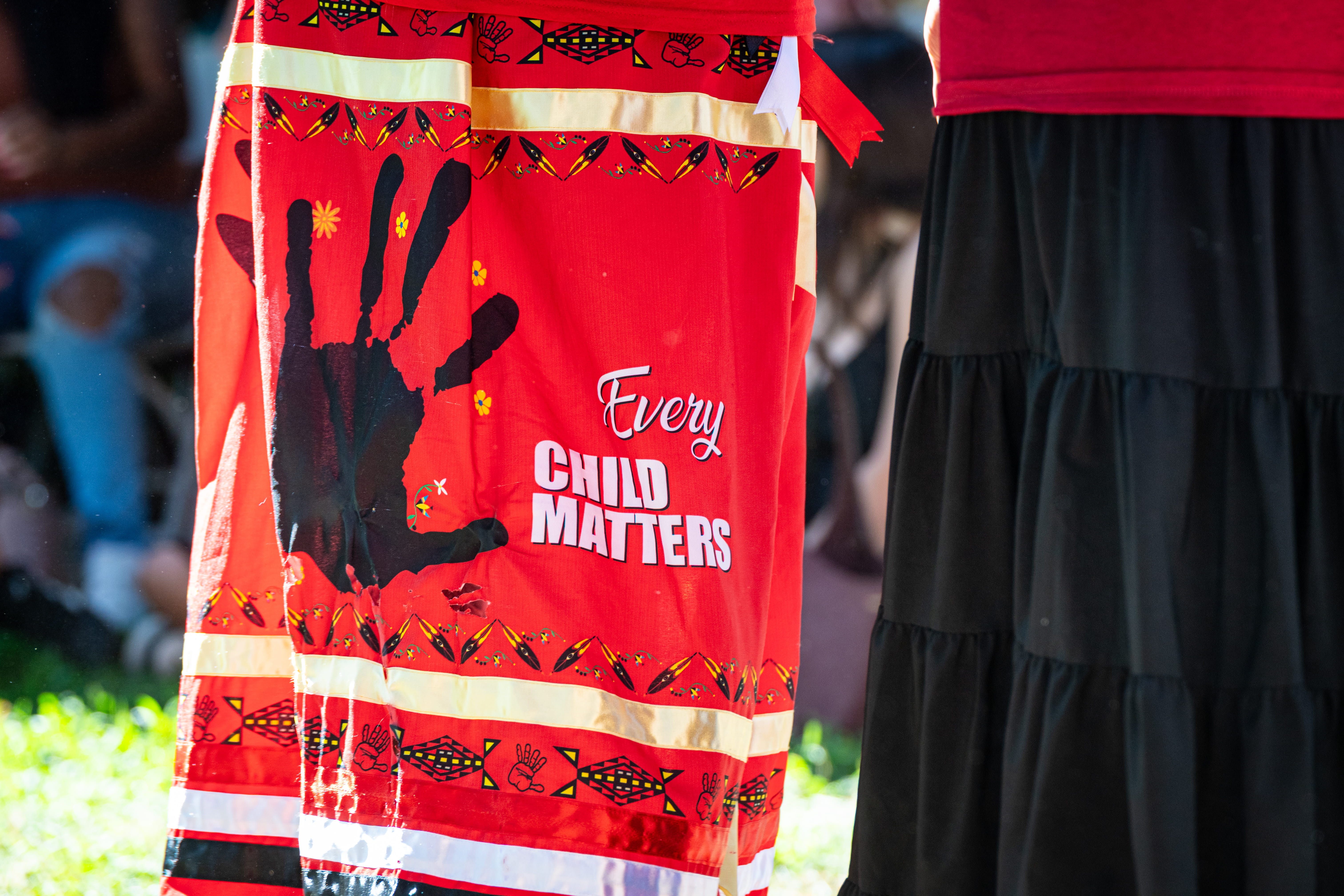 Close up of a skirt combining an Every Child Matters message with a Missing and Murdered handprint symbol on red at a public park.