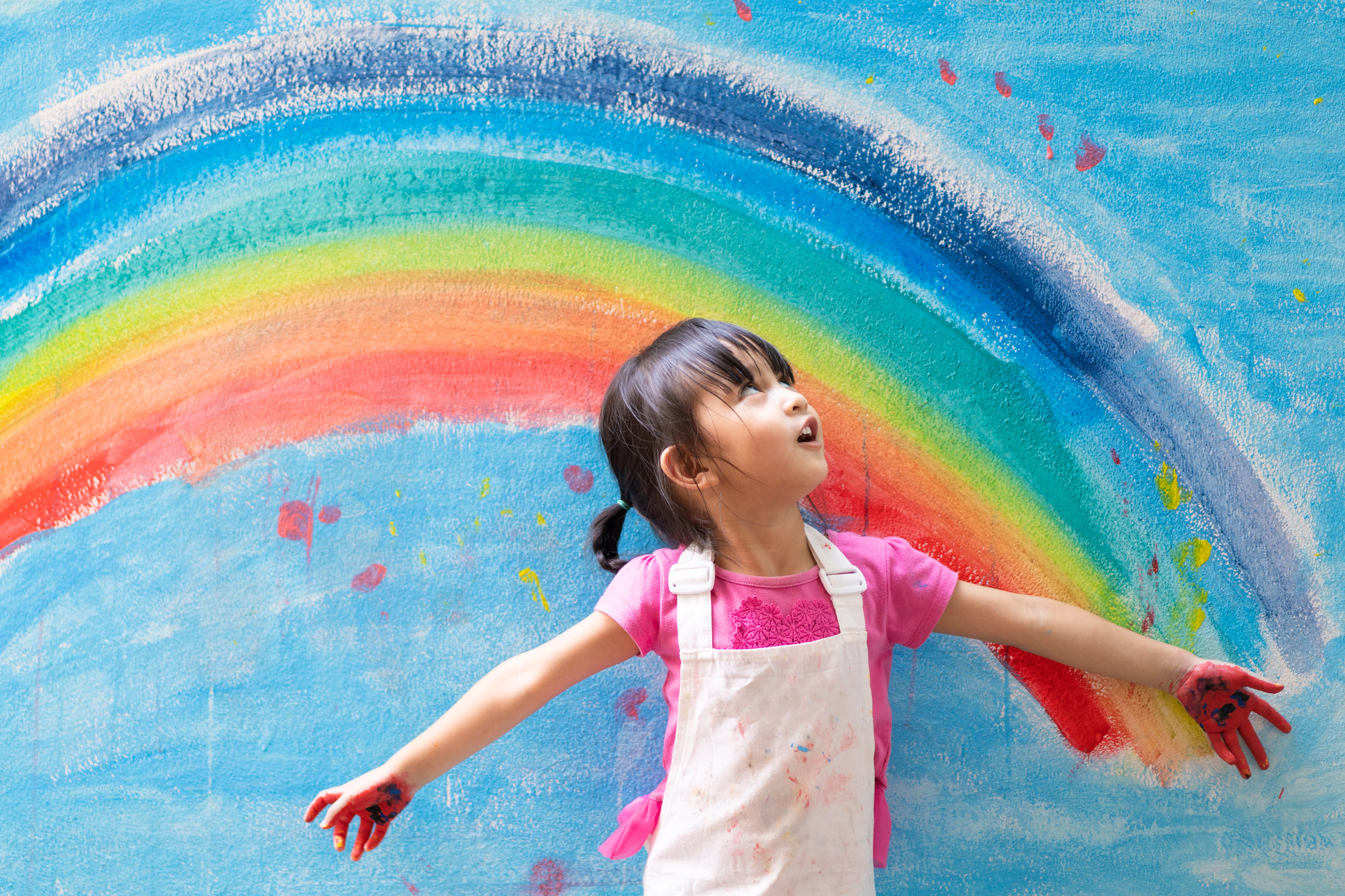 Asian little girl is painting the colorful rainbow and sky on the wall and she look happy and funny, concept of art education and learn through play activity for kid development.
