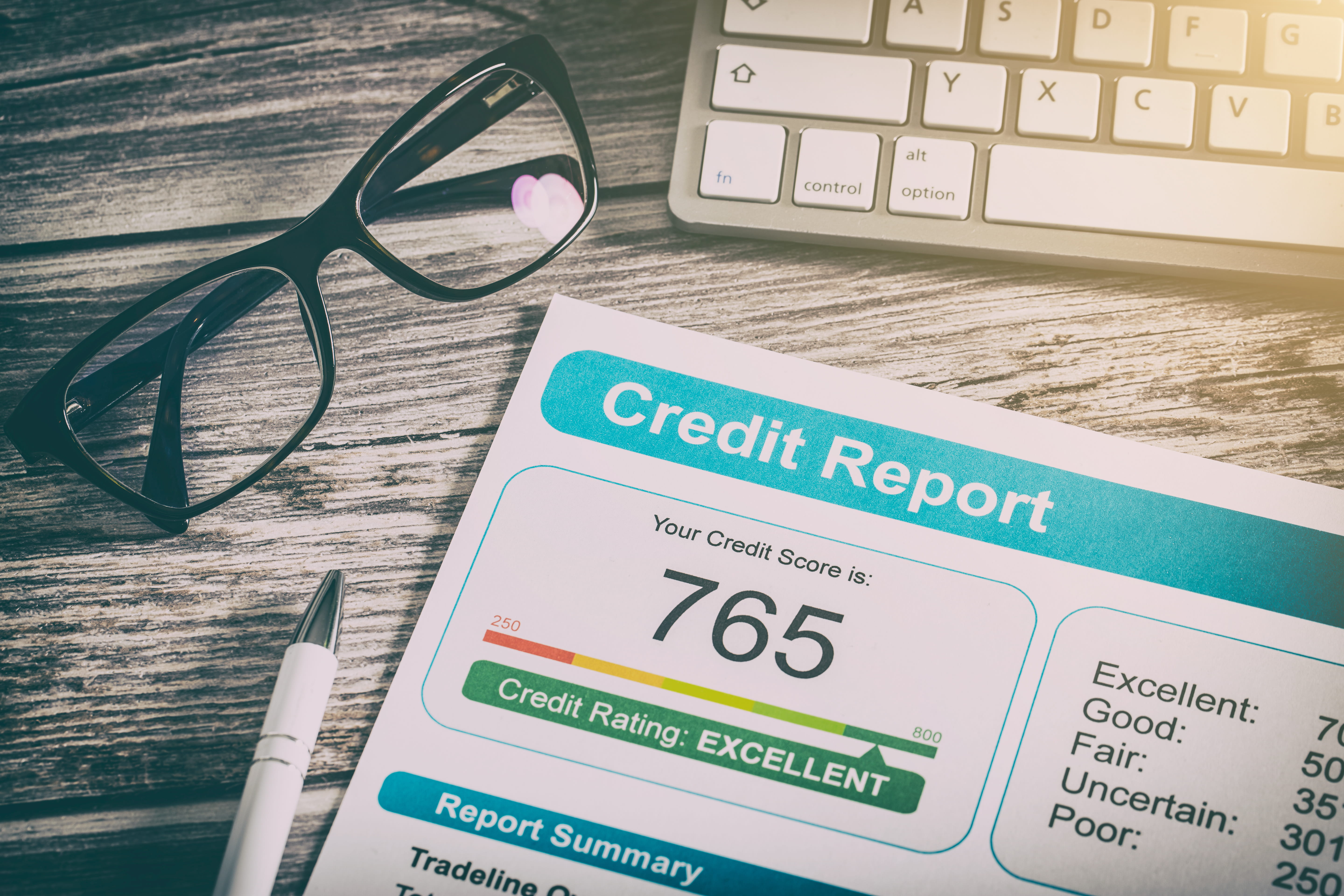 A credit report of 765 sits on a desk with a computer keyboard and glasses