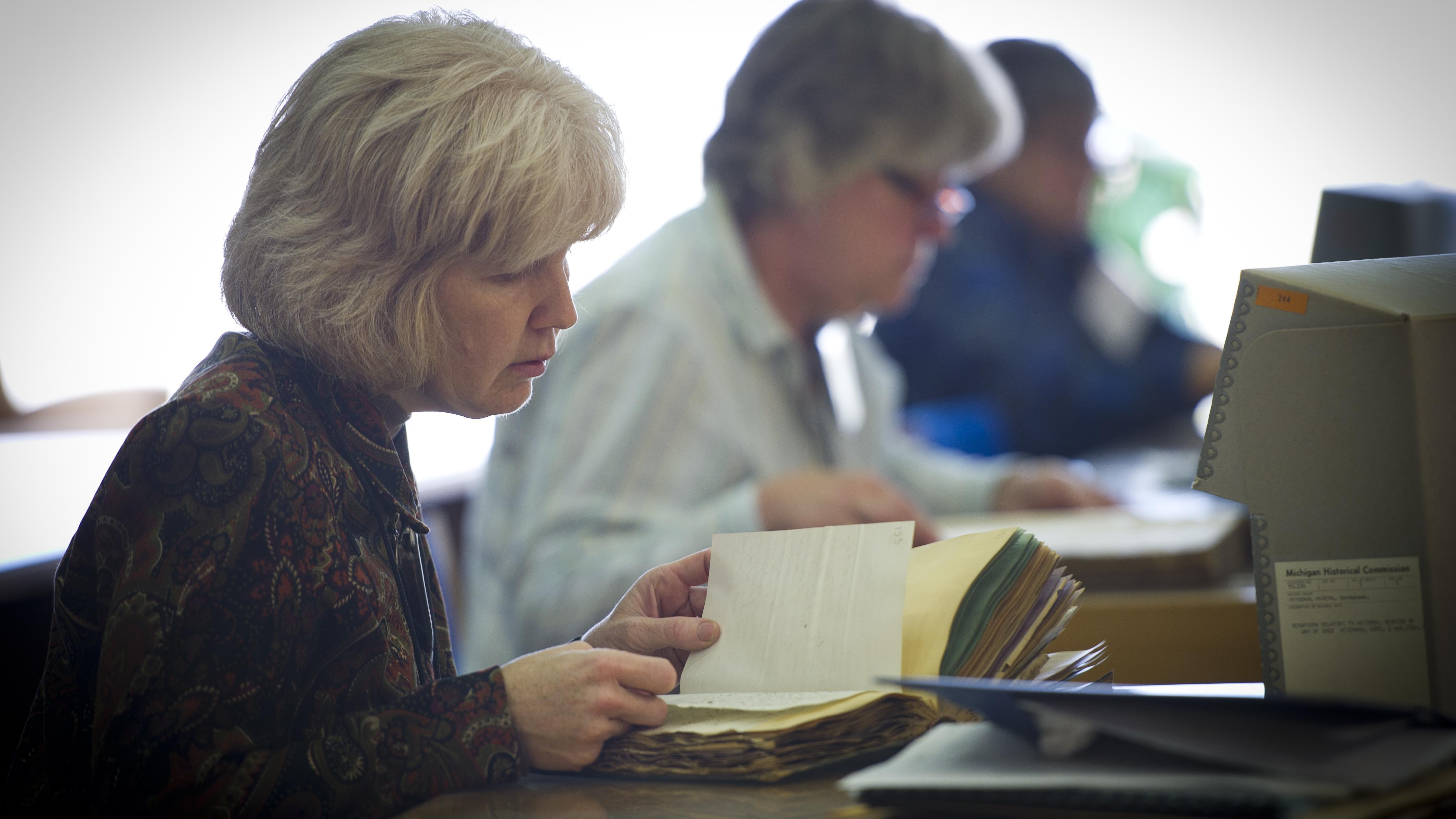 A women sitting at a table leafing through a volume. Two additional researchers are in the background.