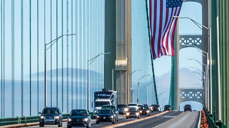 Cars driving across the Mackinac Bridge with an American Flag hanging from the bridge