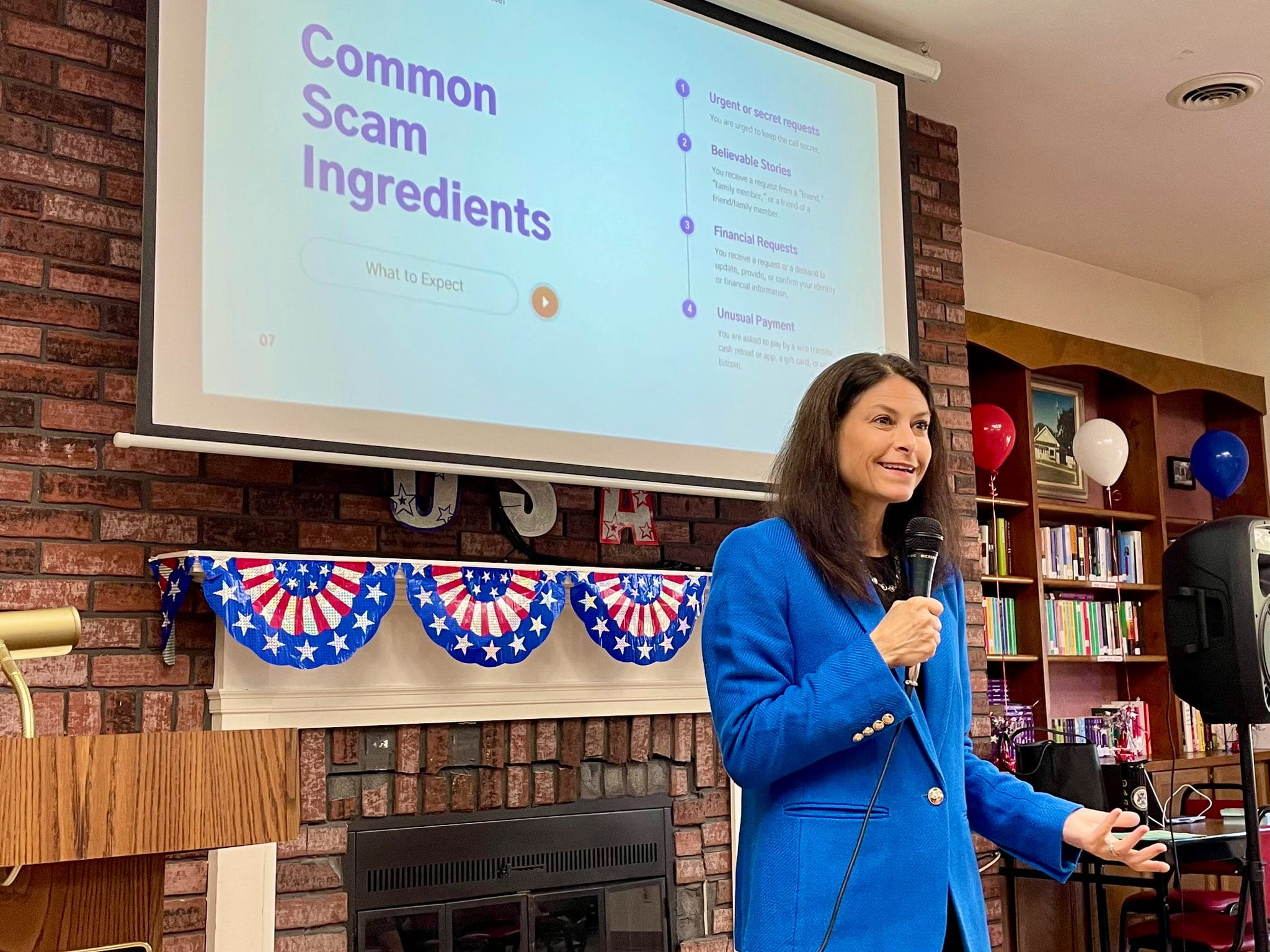 Dana Nessel giving a presentation, standing in a room in front of a fireplace next to a bookshelf, she's holding a microphone smiling.