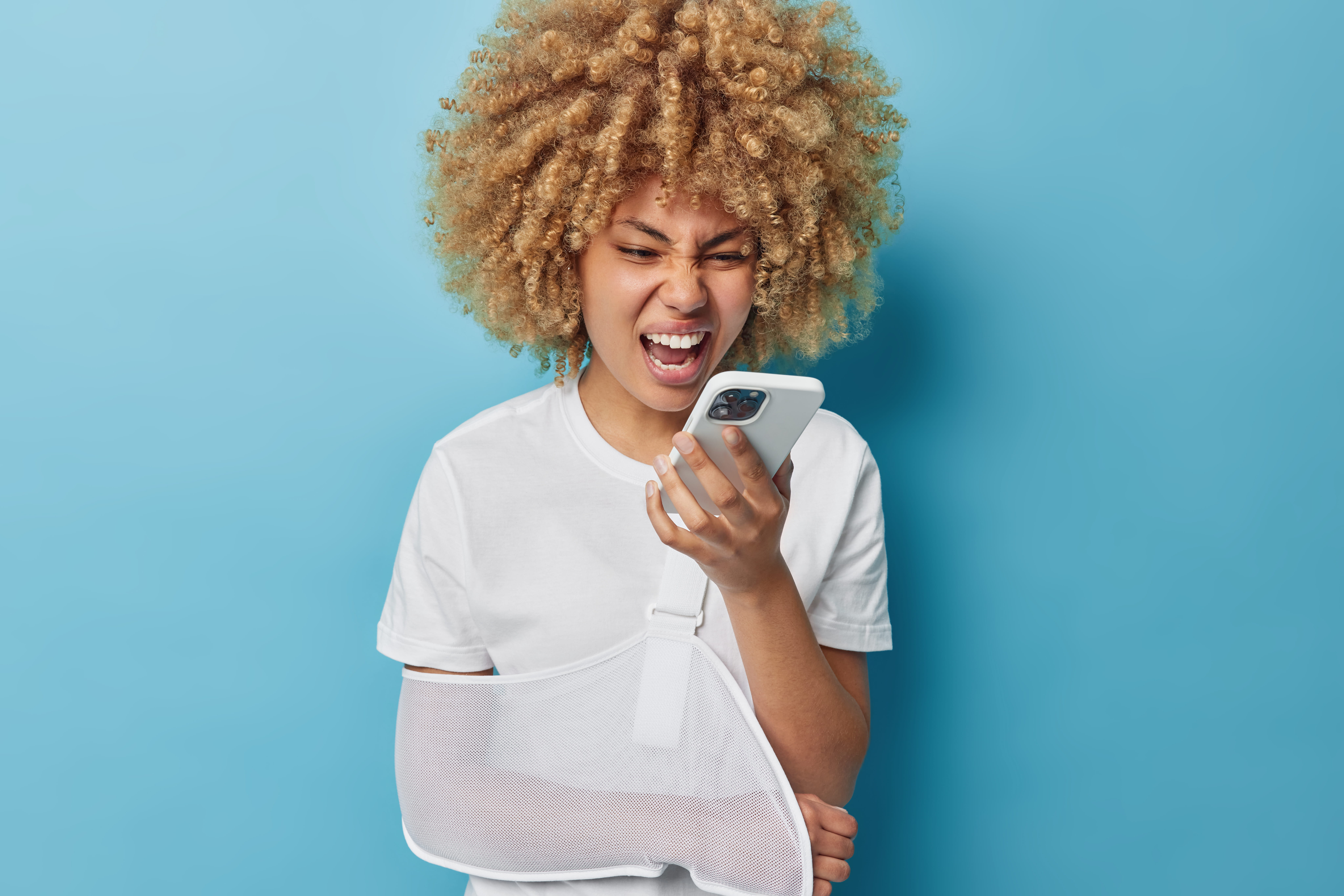 Furious woman screams in phone expresses anger shouts loudly has broken arm in sling wears casual white t shirt isolated over blue background