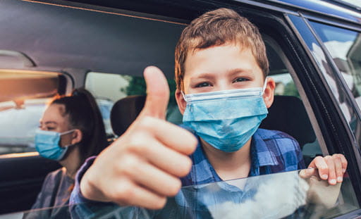 Young boy wearing a mask, looking out passenger car window with thumbs up.