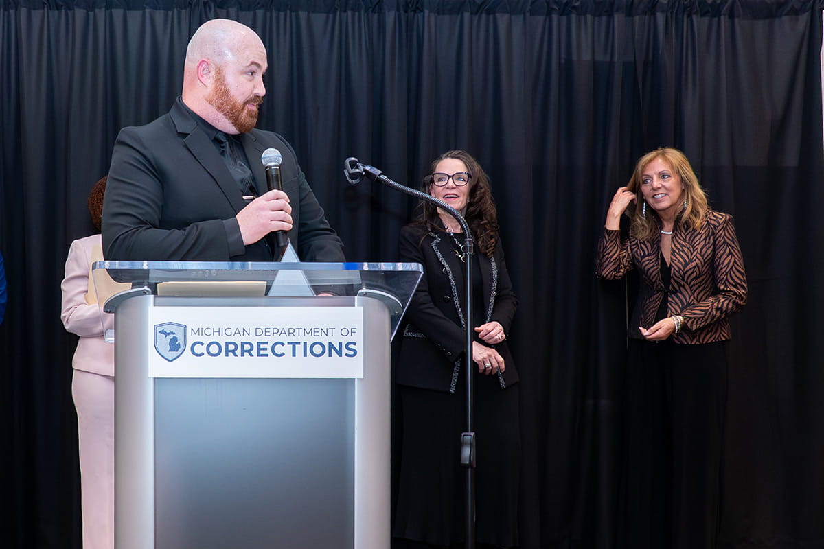 A man in a suit speaking at a podium while two women behind him, watch him speak.