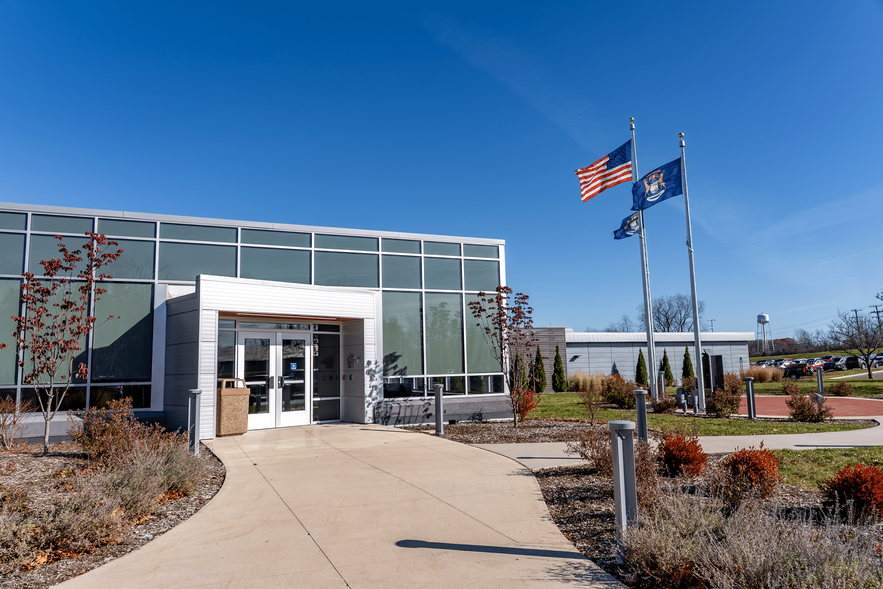The entrance to a building with a curving sidewalk leading up to the door. The USA flag and the state of Michigan flag are waving in the background on a clear day.