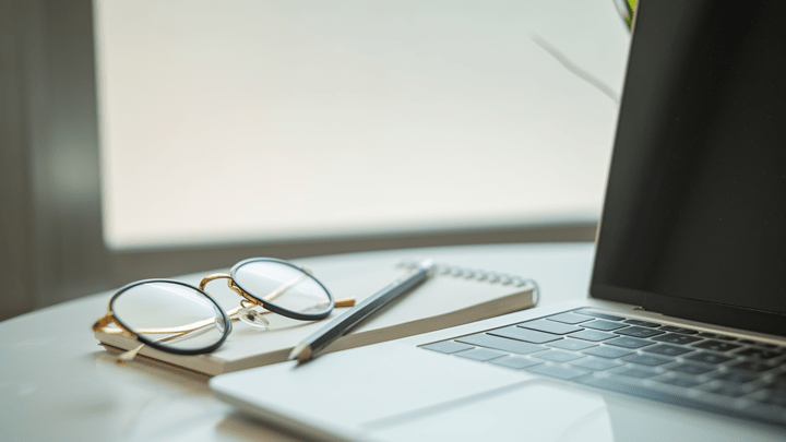 Open laptop with a notepad beside it, a pair of folded eye glasses rests on top of the notepad.