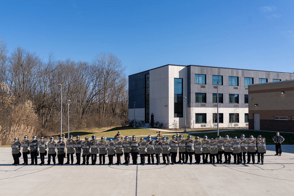 A row of corrections officer recruits holding up a safety shield. They are all outside on a sunny day and standing in an outdoor basketball court.