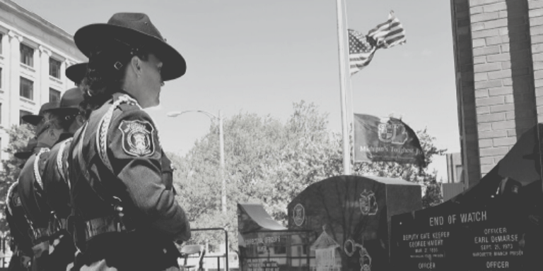 Uniformed officers standing in front of an End of Watch memorial plaque with American flag at  half staff in the background