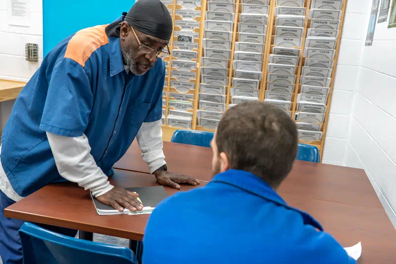 Two prisoners gathered around a table in an employment counseling office with brochures from other Michigan counties arranged on the wall behind them.