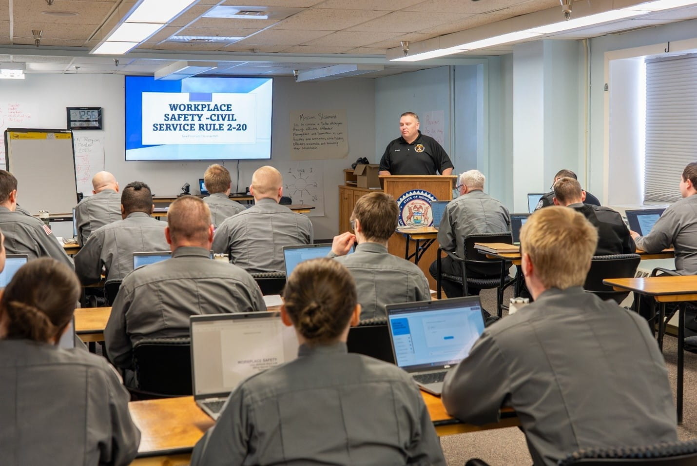 A classroom with an instructor speaking at a podium and corrections officer recruits seated at tables listening to the instructor.