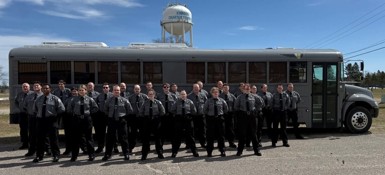 Male and female corrections officer recruits, standing at attention in front of a bus on a sunny day.