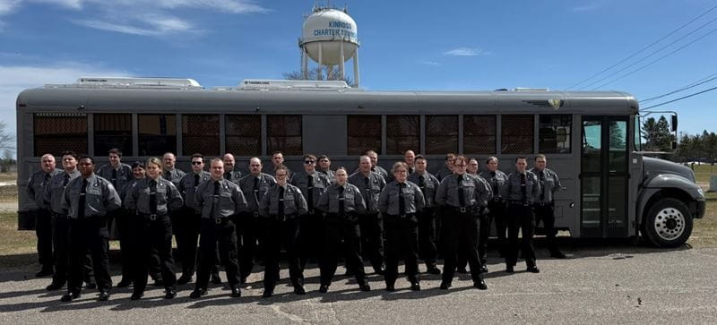 Male and female corrections officer recruits, standing at attention in front of a bus on a sunny day.