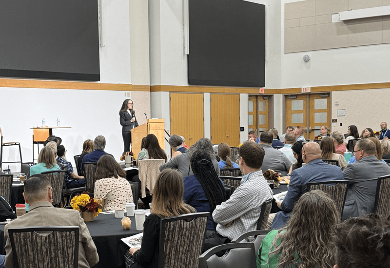 A woman standing at the front of a room and speaking to an audience of people seated at tables.