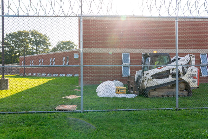 A backhoe parked on the grass behind a tall fence with razor wire.