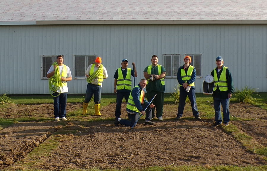 Seven male prisoners stand in a clear garden bed that is ready vegetables to grow.