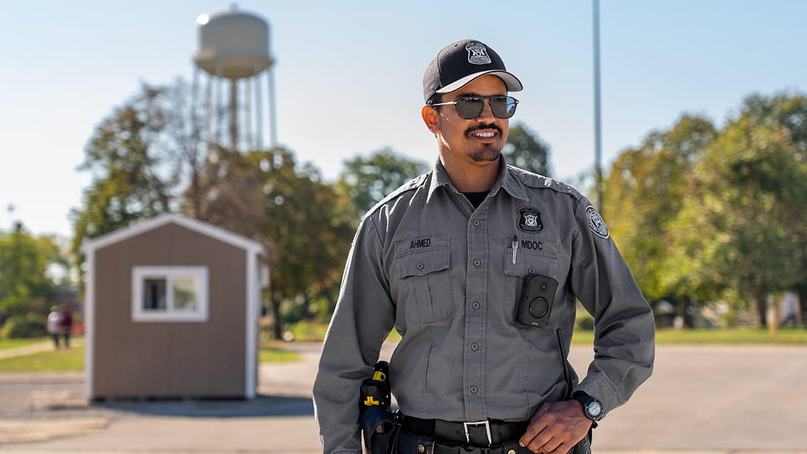 A corrections officer in his uniform standing in a prison yard with a water tower in the background.