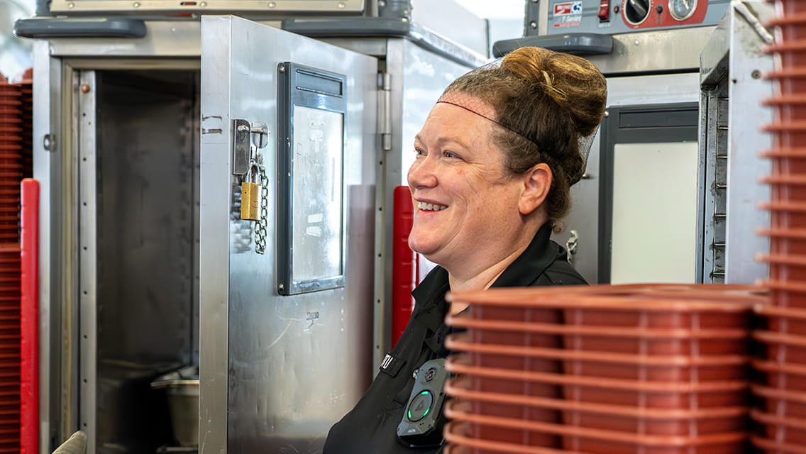 A female cook in a kitchen with a hairnet on standing behind a stack of food trays.