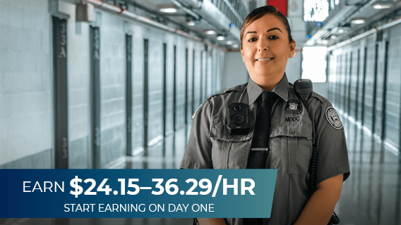 A female corrections officer standing in a hallway with holding cells on each side of her with text that reads "Earn $23.45–35.23 per hour."