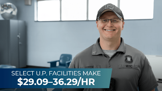 A male corrections officer standing in a prison visiting area with text that reads "Select Upper Peninsula facilities make $28.24–35.23 per hour."
