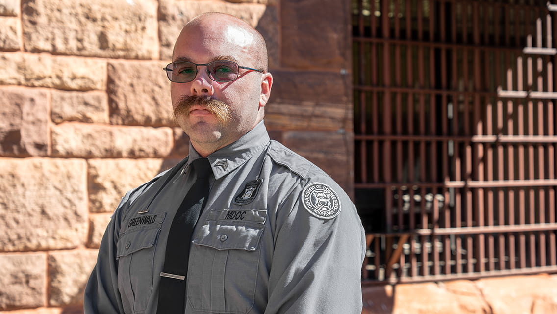 A male corrections officer in his uniform wearing glasses and standing against a brick wall.