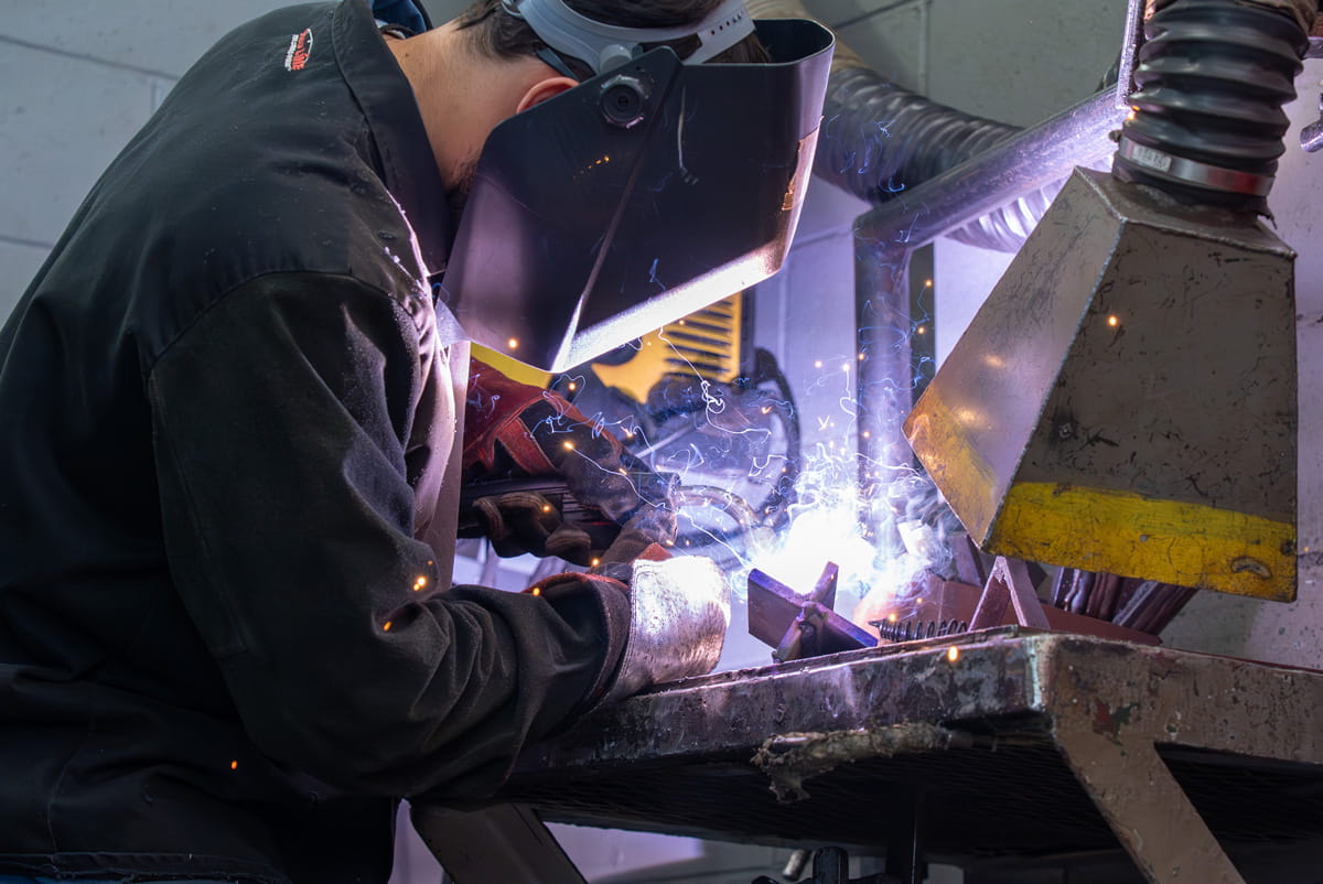 A prisoner wearing a welding visor and gloves while welding metal on a machine.