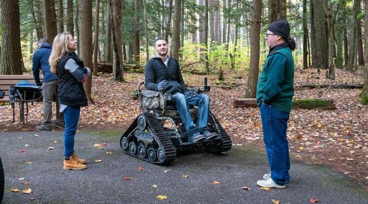 Group of three people, one in a track chair/wheelchair talking on a trail with fall leaves in background