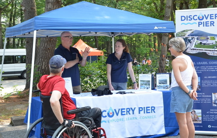 Group of people, one using a wheelchair, gathered in a group by a canopy tent talking