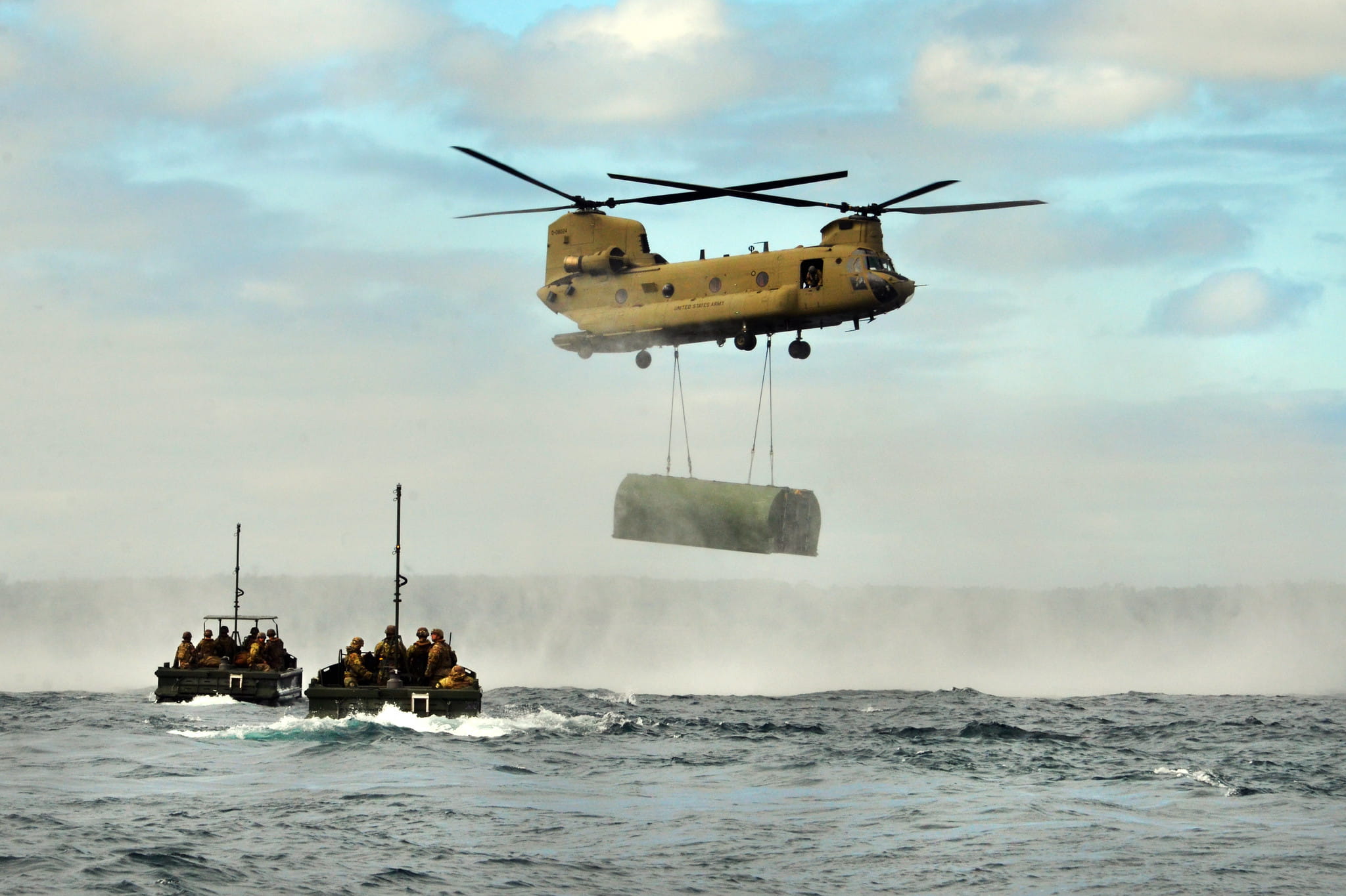 chinook helicopters carry bridging equipment over water while two boats observe