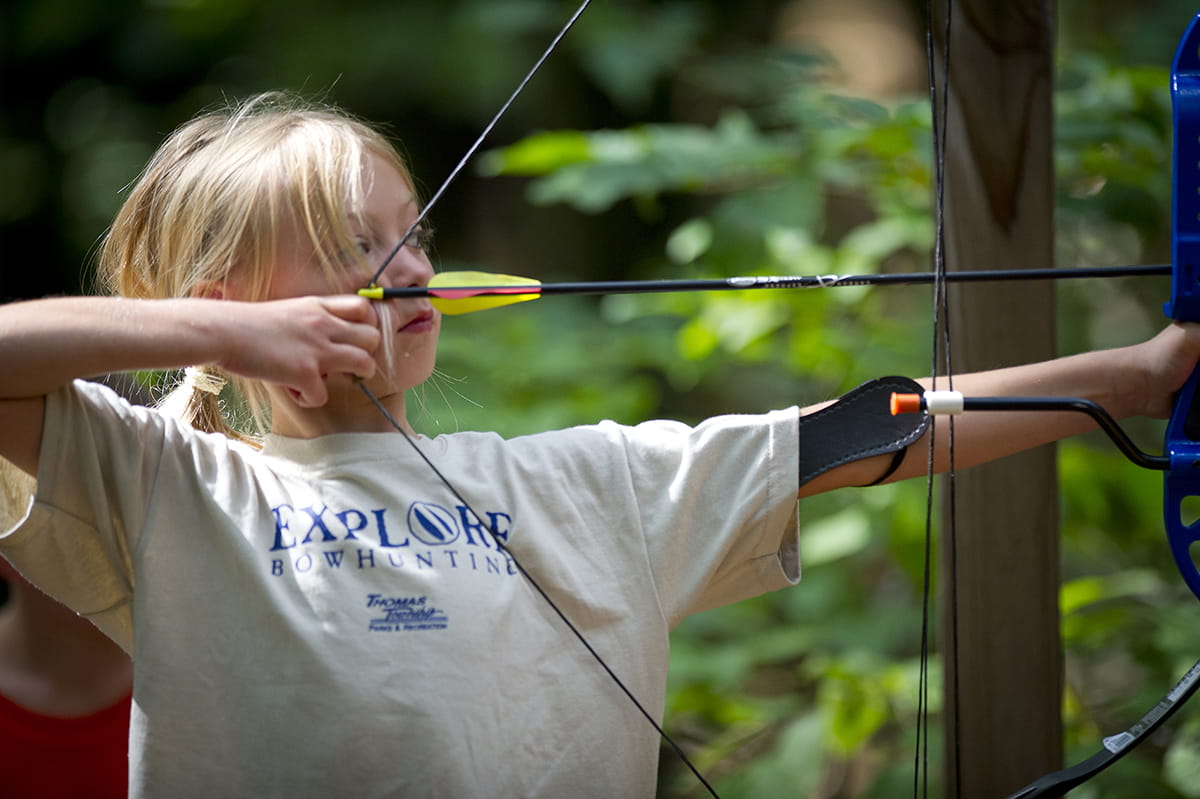 Young girl wearing Explore Bowhunting shirt pulls back an arrow preparing to shoot a bow