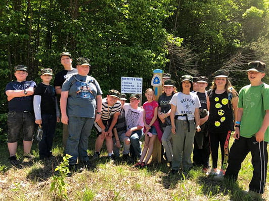 Group of boys and girls standing in the grass on a trail with woods in the background