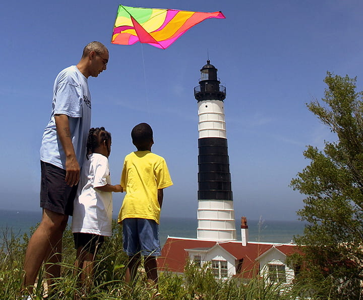 A man and two children fly a kite in front of the Big Sable Lighthouse