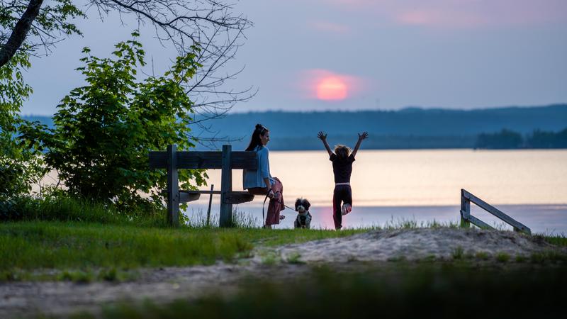 A woman sits on a bench watching a sunset over a lake with two children, one has arms up in the air while jumping