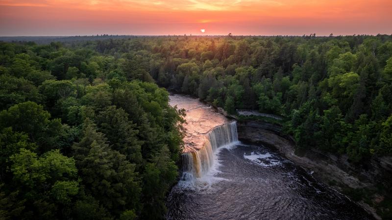 Aerial view of a large waterfall from a river going through the trees with a vibrant sunset in the sky. The sunset colors reflect off the water as it falls into the pool below.