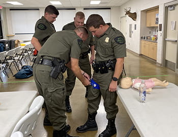 A man applies a tourniquet to a man standing in a classroom