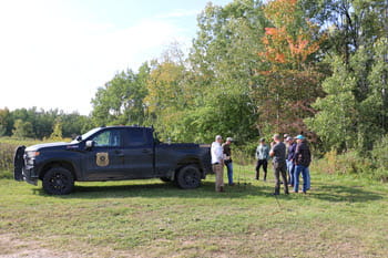 A group of people stand near the woods behind a CO patrol truck