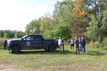 A group of people stand near the woods behind a CO patrol truck