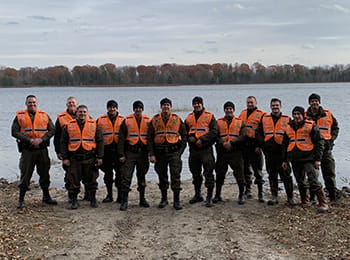 12 officers wearing orange safety vests stand smiling by a lake