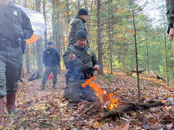 man kneeling on ground in the woods puts a stick in a fire