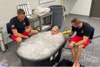 Two lifeguards sit next to a man who is sitting in a tub full of ice.