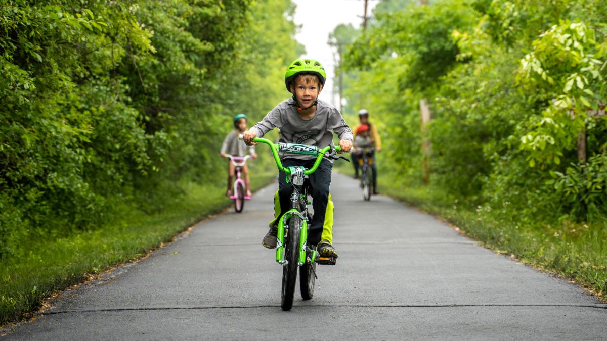 Three children ride bikes down a paved path flanked by green trees