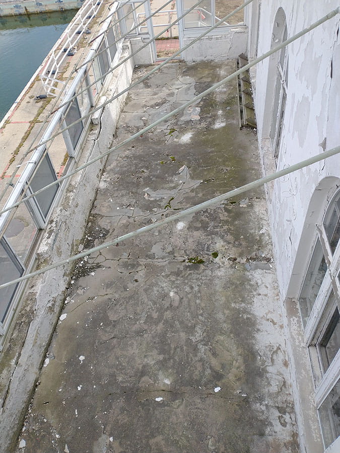 Exterior porch with roof missing. Concrete on the floor has cracks and vegetation growing in cracks.