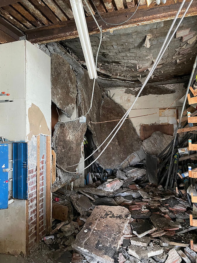 Room with collapsing ceiling with wires hanging down from it. Floor filled with concrete debris.