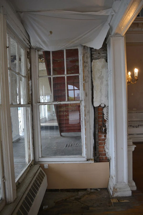 Room with paint chipping on the two window boarders. Wall adjacent is deteriorating with ceiling collapsing.  