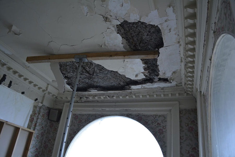 Exposed deteriorated concrete ceiling with missing pieces held up with a wooden support beam.