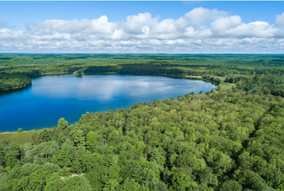 An aerial view of a lake surrounded by green forest with blue sky and clouds overhead.