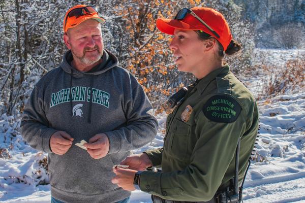 A conservation officer talks with a hunter out in the field. Both wear hunter orange hats.