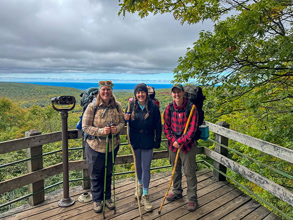 three backpackers stand on platform overlooking forest and lake at the Porkies